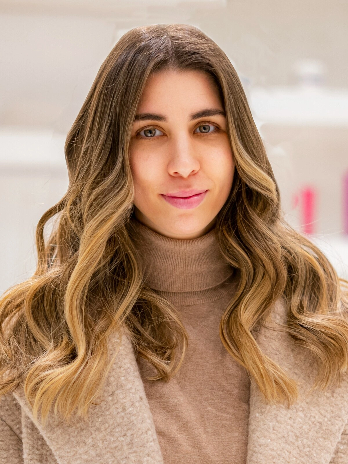 A woman wearing a camel-coloured roll-neck and wool coat with a bouncy, wavy balayage showing brunette hair blending into caramel tones