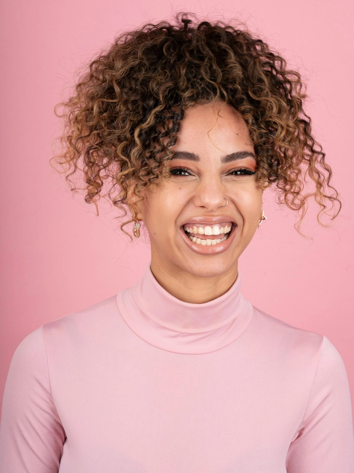 Studio shot of smiling woman with afro balayage hair wearing a pale pink turtle-neck top