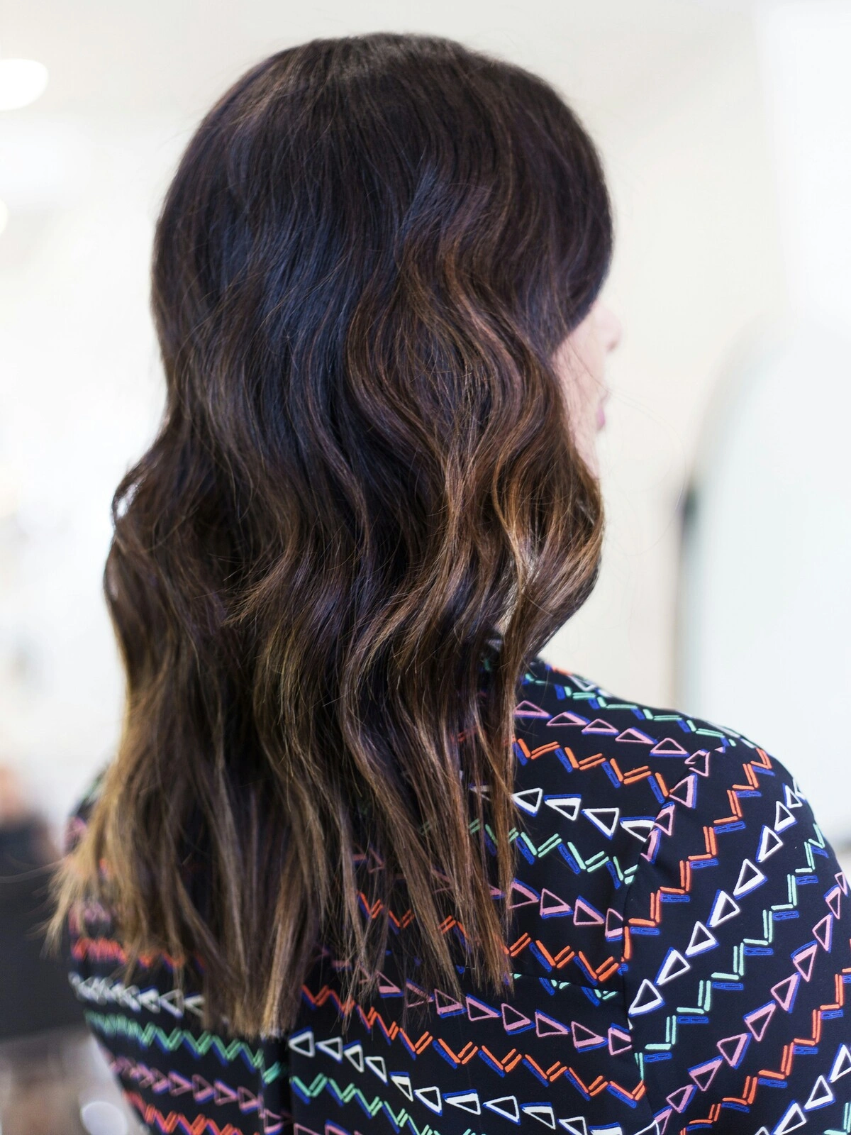 Back view of a woman’s head showing a balayage in loose waves with black hair blending into warm brunette tones