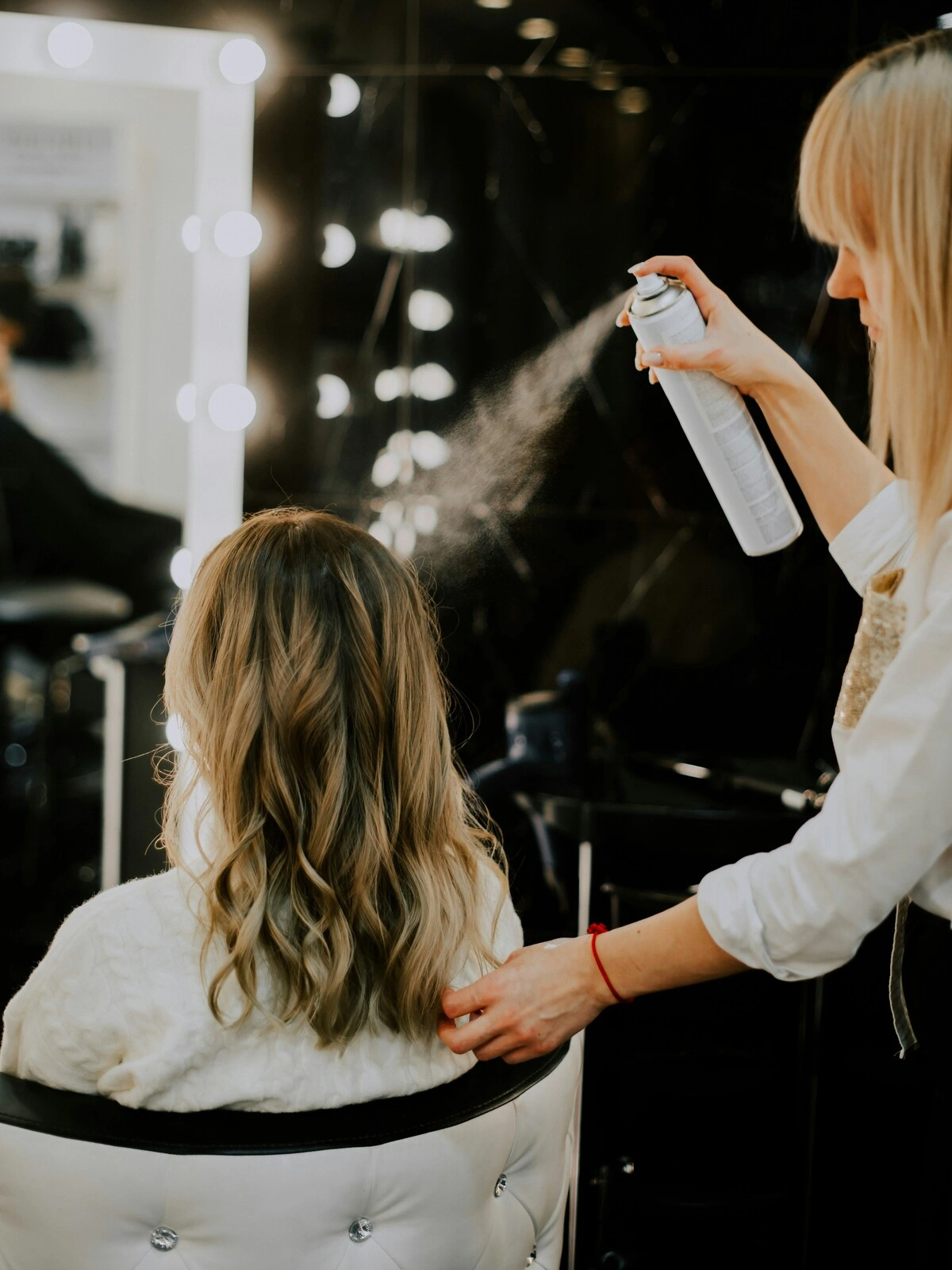 A hairdresser sprays a finishing product on a client’s hair after a balayage treatment, a blow dry and styling into waves