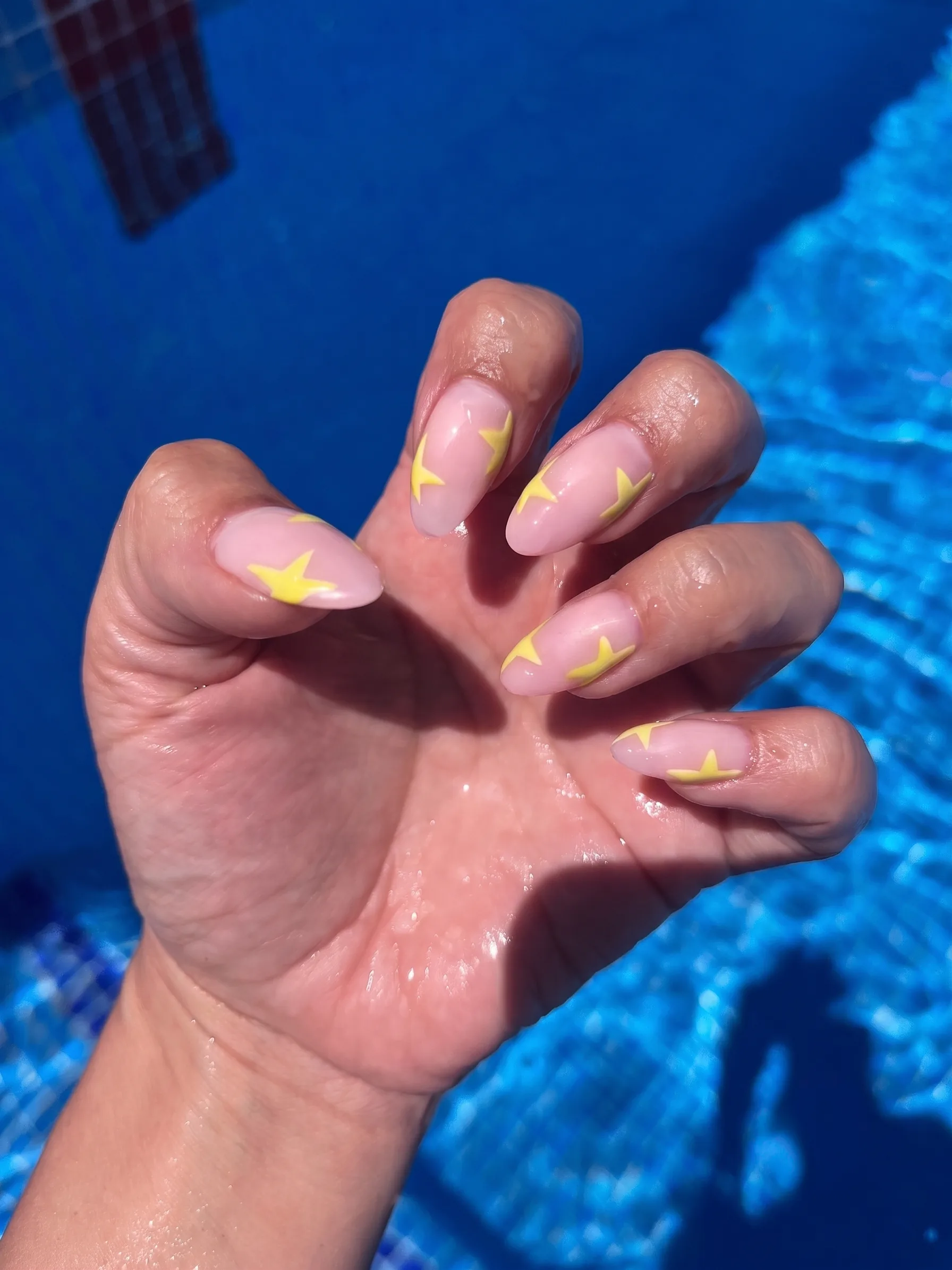 Sheer nude almond nails with butter-yellow star accents, photographed poolside