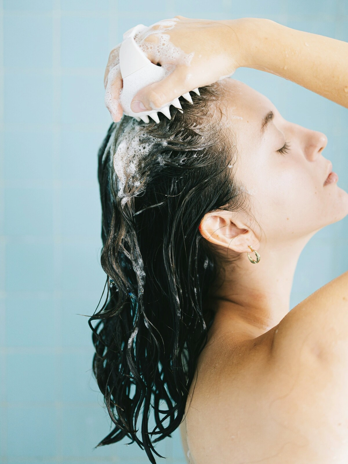 Side-view of a woman with wet shampoo hair massaging her scalp with a silicone brush