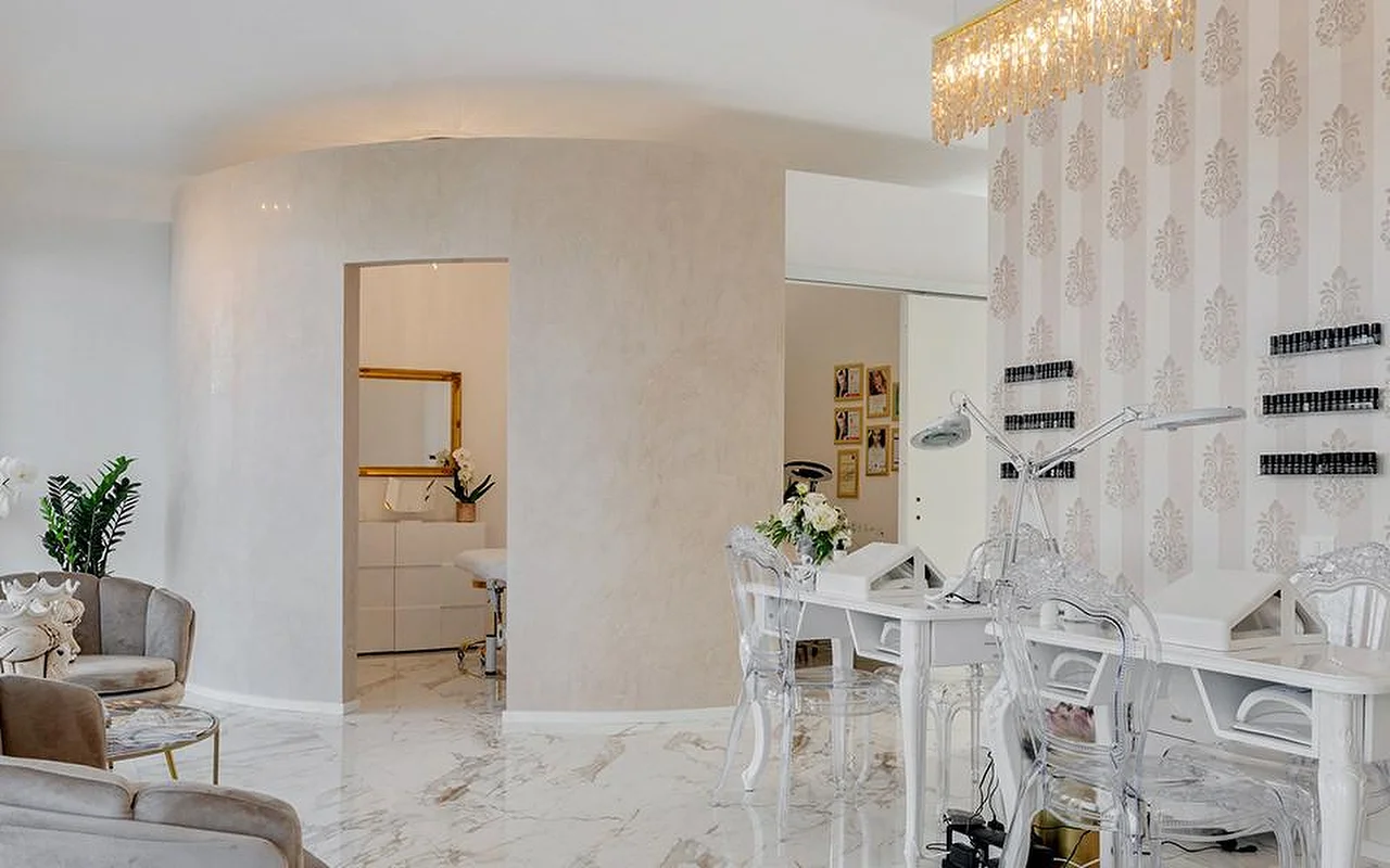 The elegant interior of MC Beauty Center with marble-effect flooring, white curved walls, and a gold crystal chandelier. A nail station with ghost chairs sits to the right, while velvet armchairs and a gold-framed doorway lead to a treatment room beyond.