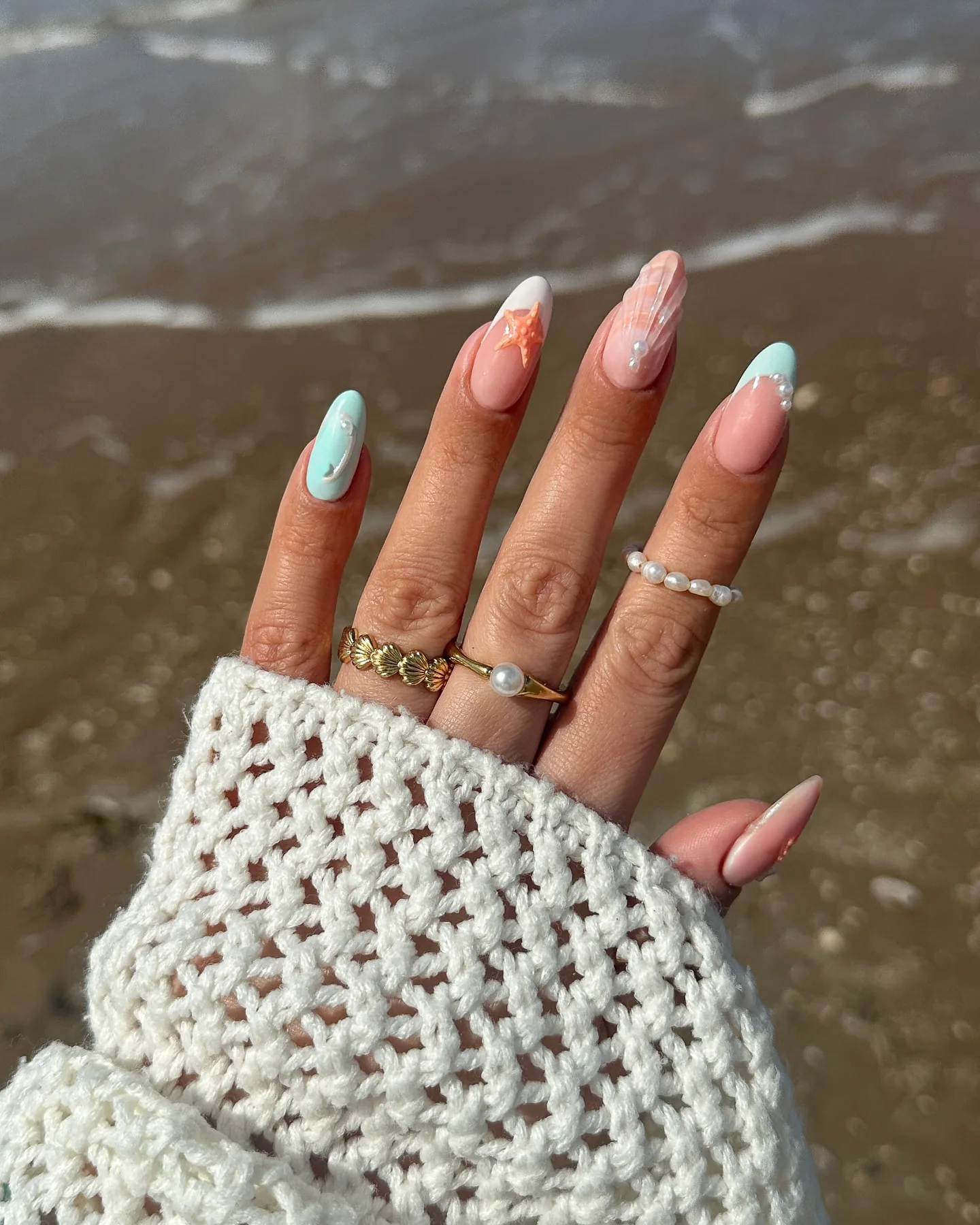 Almond nails with pastel mermaid-inspired art — a starfish, pearl accents and pearly shimmer — held against wet sand