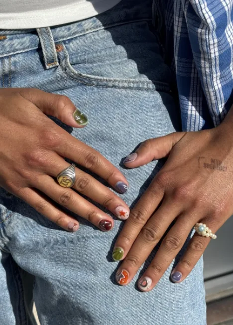 Short mismatched manicure with each nail a different colour and design, including a red star and an orange swirl, resting on blue jeans