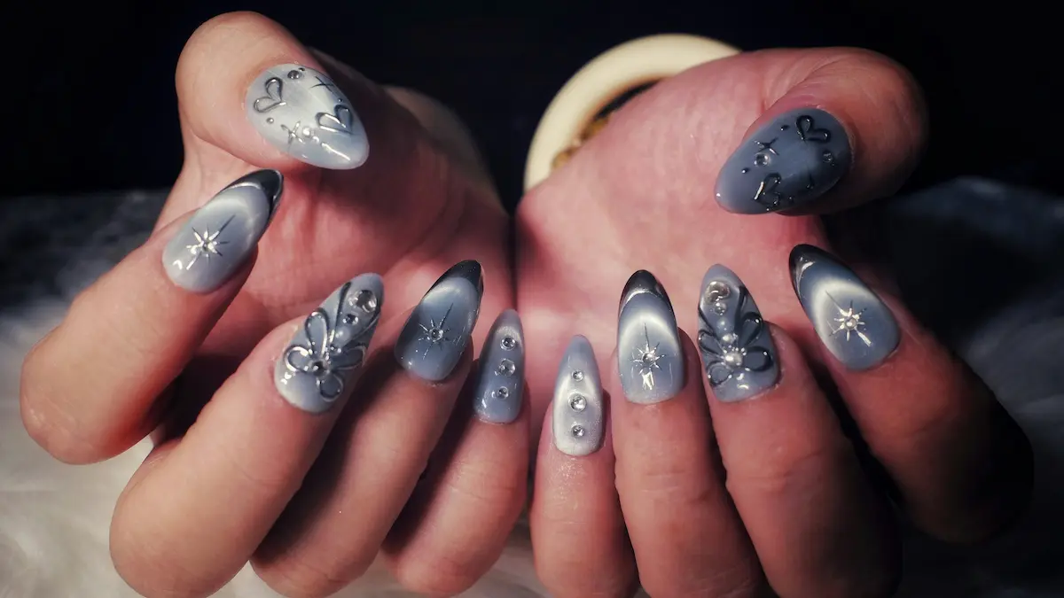 Close-up of a woman’s hands showing intricate acrylic nails with a grey cats-eye design, silver hearts and stars, and sparkling gemstone embellishments