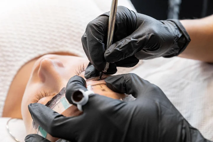 A close-up of an eyebrow treatment in progress, a technician in black gloves maps and marks a reclining client's brow with a measuring tool and pencil, the client's eyes closed.
