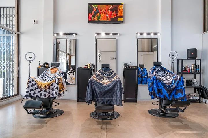 A bright, high-ceilinged barbershop with three black barber chairs draped in patterned capes, each facing a mirror framed by light bulbs, with ring lights, a wall-mounted screen and tall windows.