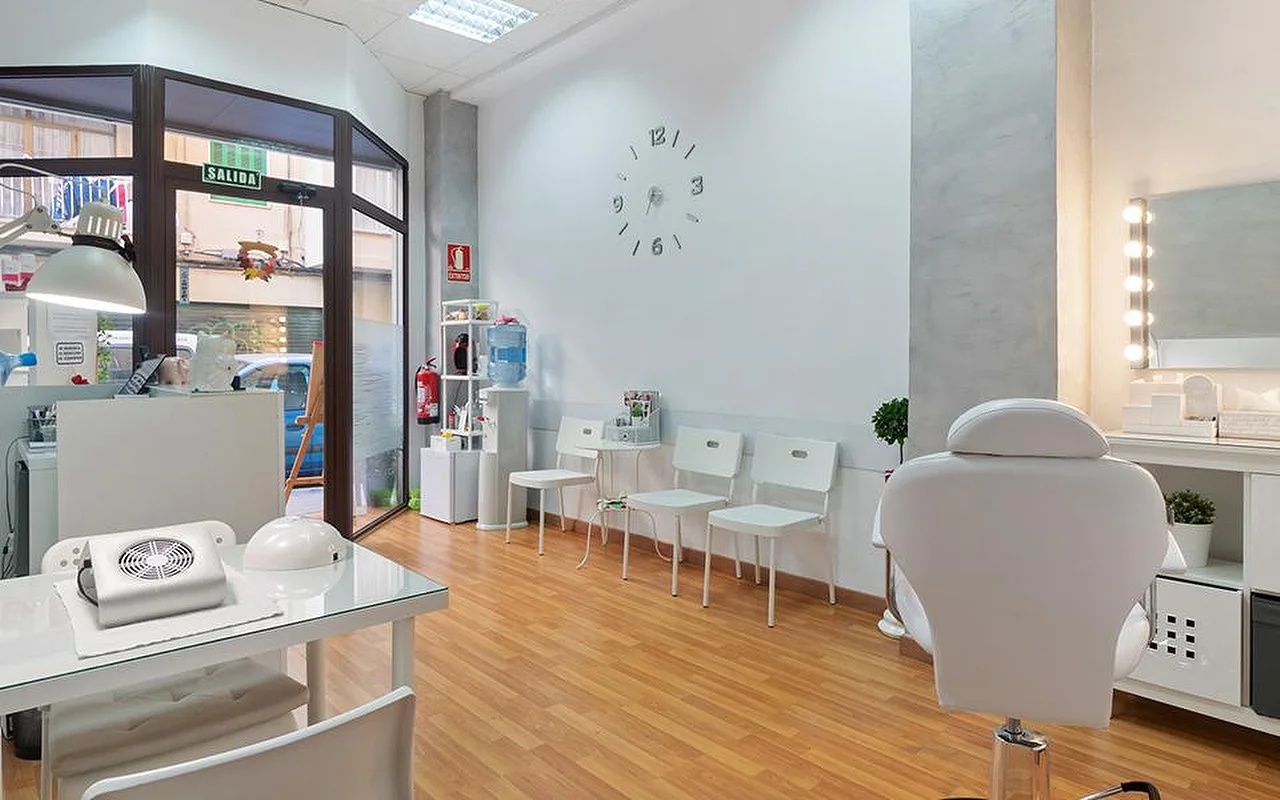 A bright, white salon with pale wood flooring, a row of white waiting chairs, a minimalist wall clock, an illuminated dressing-table mirror, a white reclining treatment chair, and a manicure station by the window.