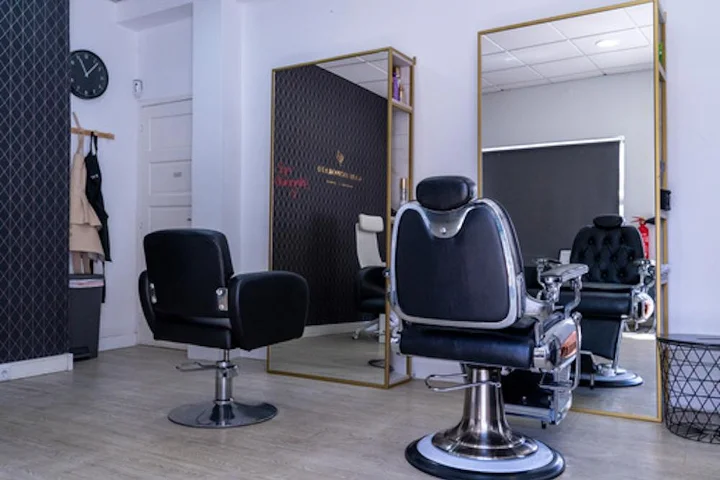 A pared-back salon interior with a black styling chair and a chrome-and-leather barber chair on pale wood flooring, a large gold-framed mirror, a wall clock and aprons hanging beside a dark feature wall.