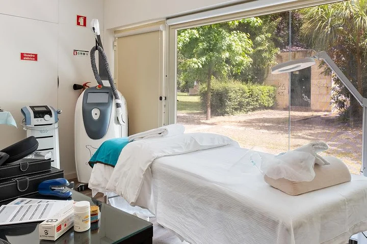 A beauty treatment room with a white-draped therapy bed beside a floor-to-ceiling window onto a green garden, a body-treatment machine on a stand, a magnifying lamp and a glass side table with products.