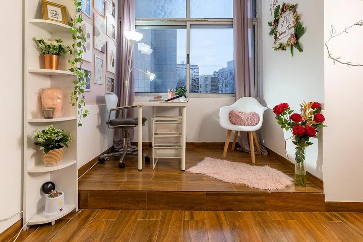A small aesthetics studio on a raised wooden platform, with a white desk and clip lamp, an office chair and a tub chair, corner shelves of plants, a vase of red roses and a window onto the city.
