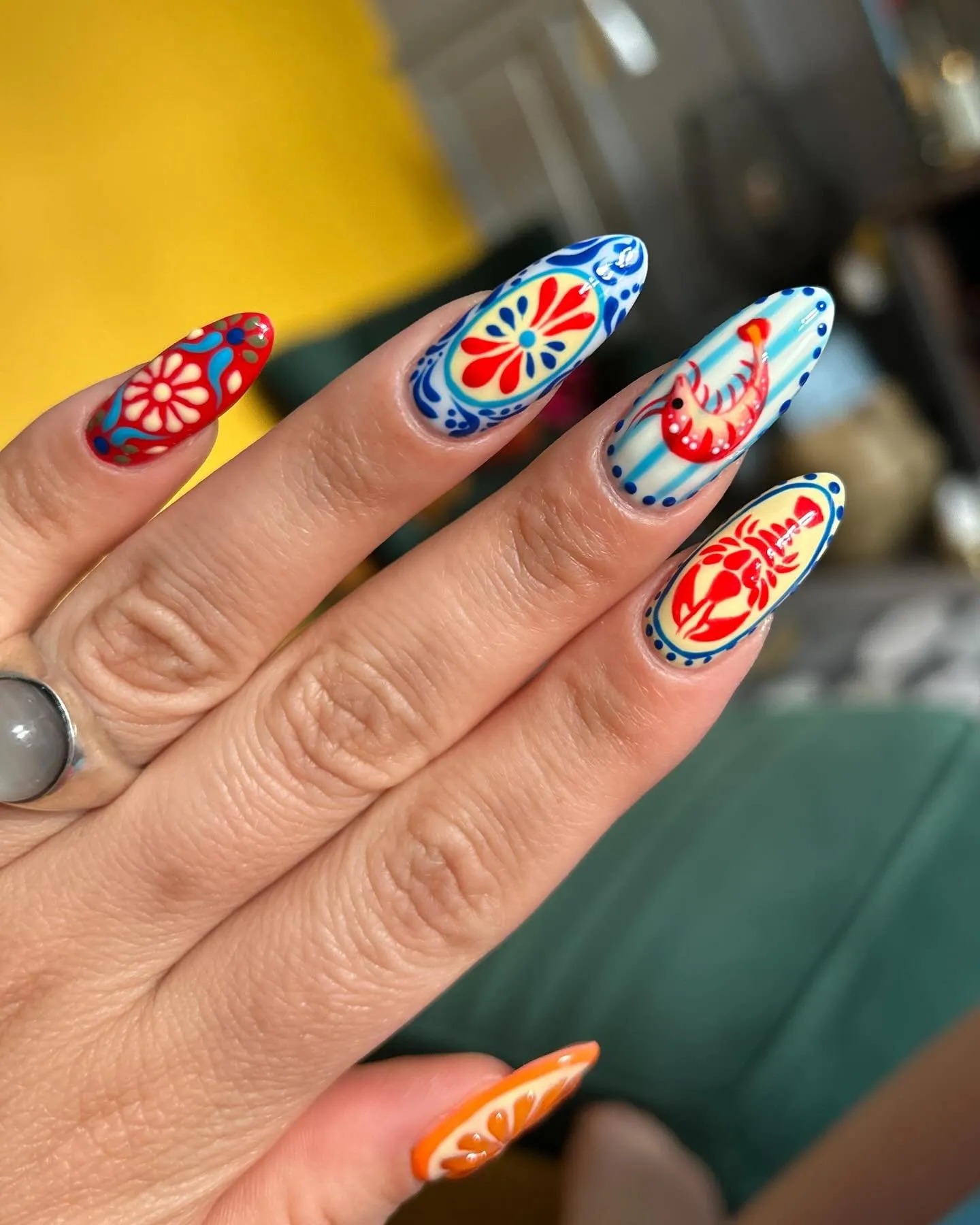 Destination nail art on almond-shaped nails featuring Mediterranean-inspired motifs in red, blue, and orange
