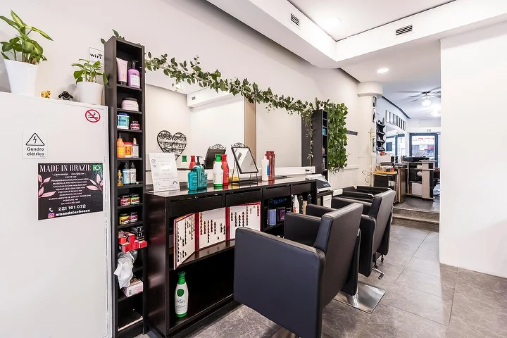 A narrow hair salon with black styling chairs along a mirrored wall, a dark shelf stacked with haircare products, a trailing greenery garland over the mirror and a framed Made In Brazil sign on a white column.