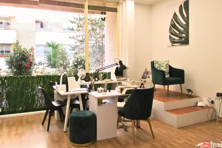 A bright manicure area with two white nail tables and lamps, green velvet chairs and a matching stool, a monstera-leaf canvas on the wall and large windows onto a planted terrace.