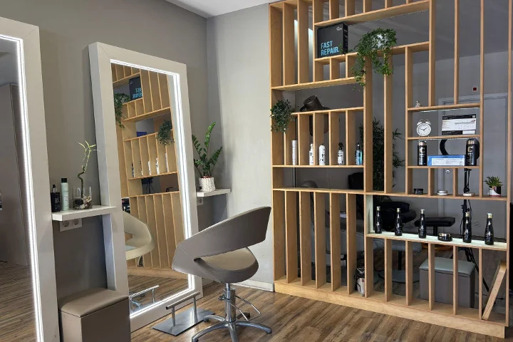 A hair station with a grey styling chair facing an LED-lit framed mirror, a wood-slatted shelving partition stocked with styling products, a clock and plants, on wood-effect flooring.