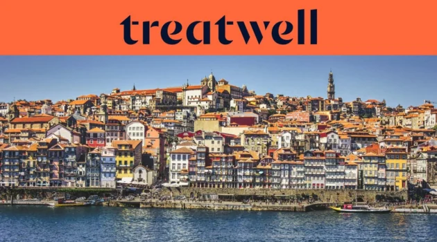The Ribeira waterfront in Porto, with rows of colourful houses and terracotta rooftops rising up the hillside from the Douro river and the Clérigos tower on the skyline under a clear blue sky.