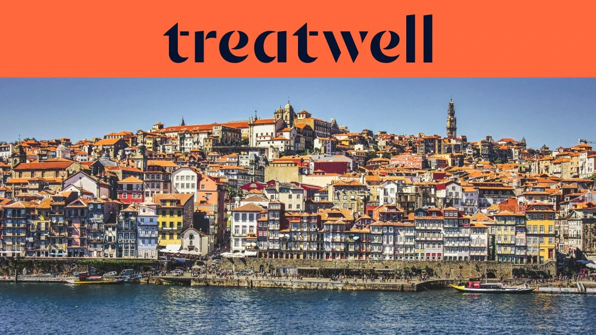 The Ribeira waterfront in Porto, with rows of colourful houses and terracotta rooftops rising up the hillside from the Douro river and the Clérigos tower on the skyline under a clear blue sky.