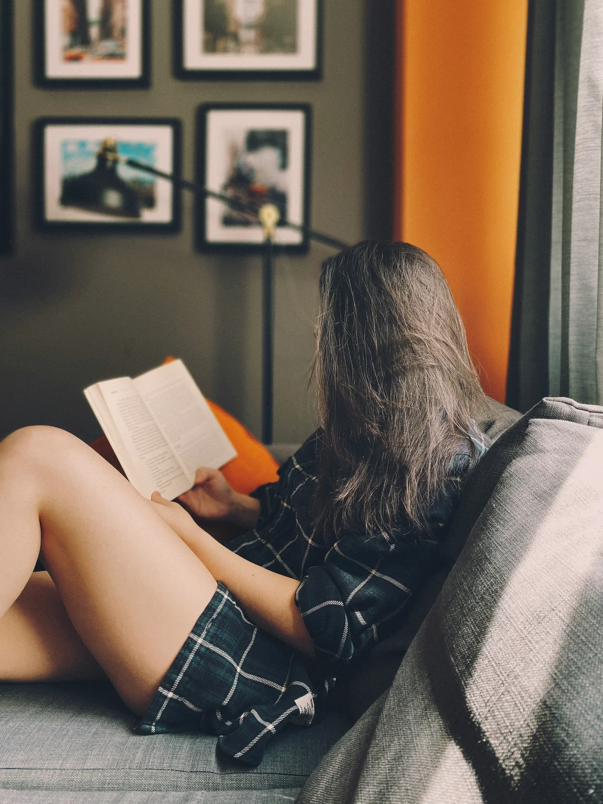 Side view of a woman sitting on a sofa reading a book with her long hair obscuring her face, she wears loose cotton shorts and a shirt in dark green check