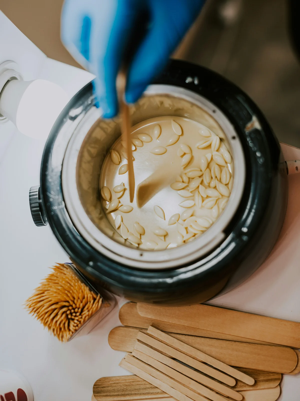 Top view of hot wax beads melting in a waxing pot with a blue-gloved hand stirring the wax with a spatula