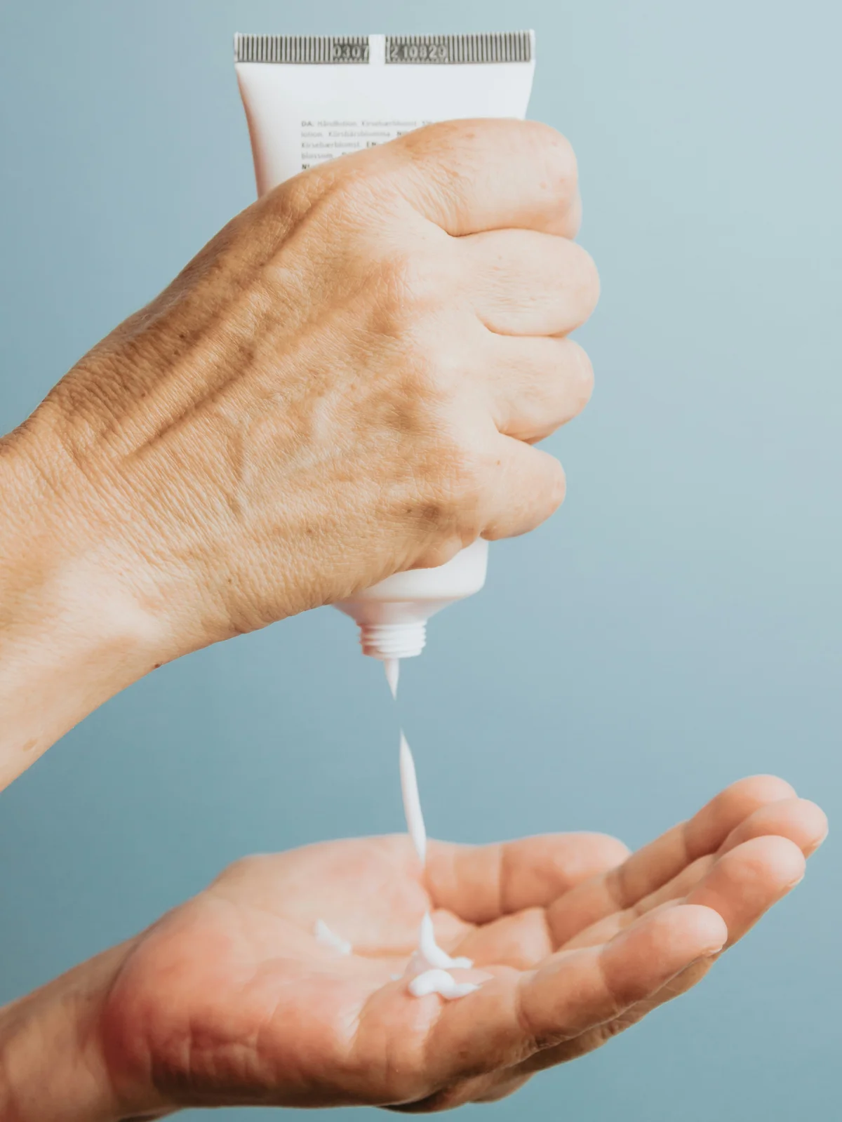 Close up of a hand holding a tube of white moisturiser upright and squeezing some into another hand underneath