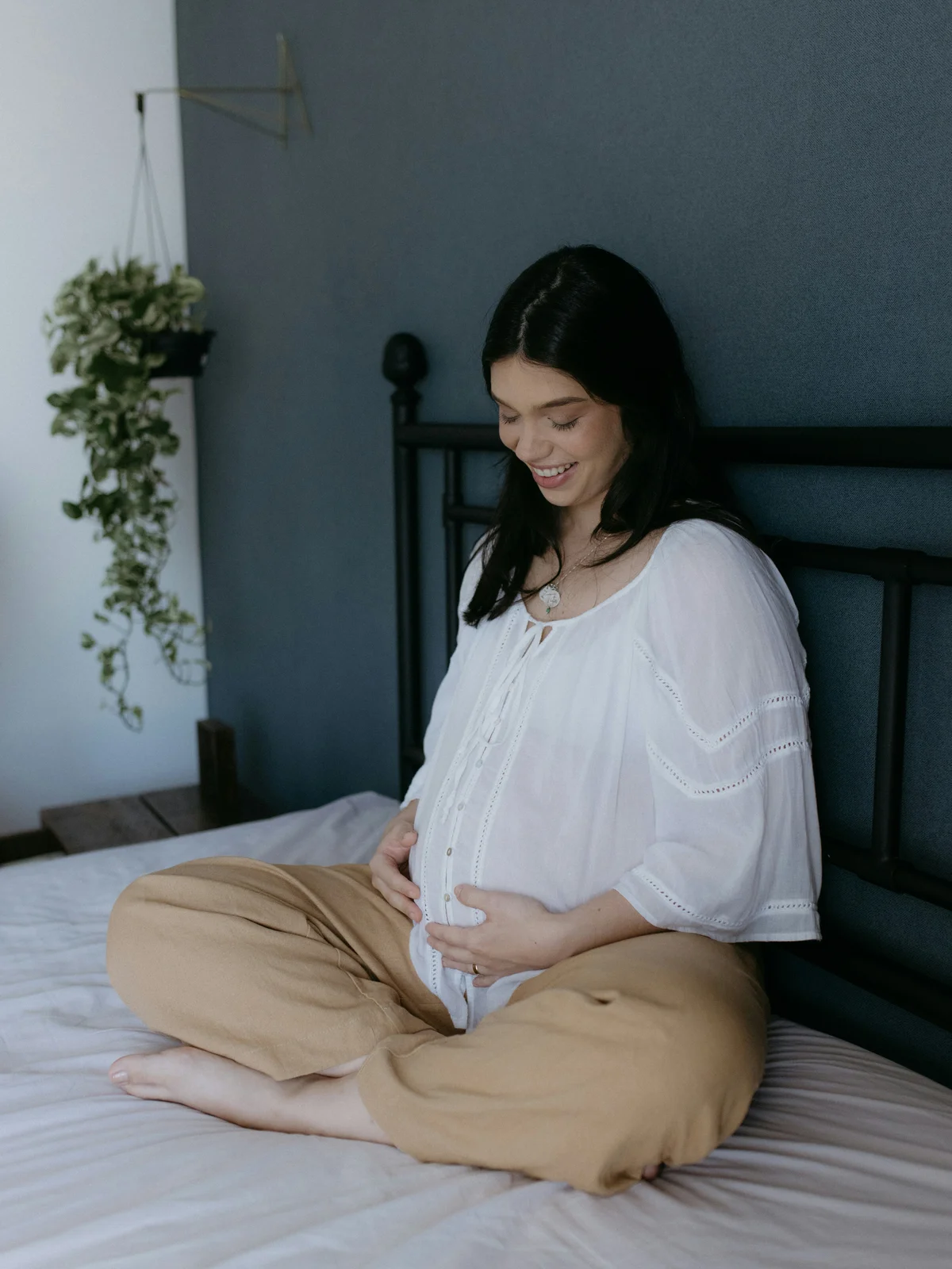 A pregnant woman sits on a bed wearing a white shirt and beige trousers with her legs crossed, hands on her bump, and smiling downwards