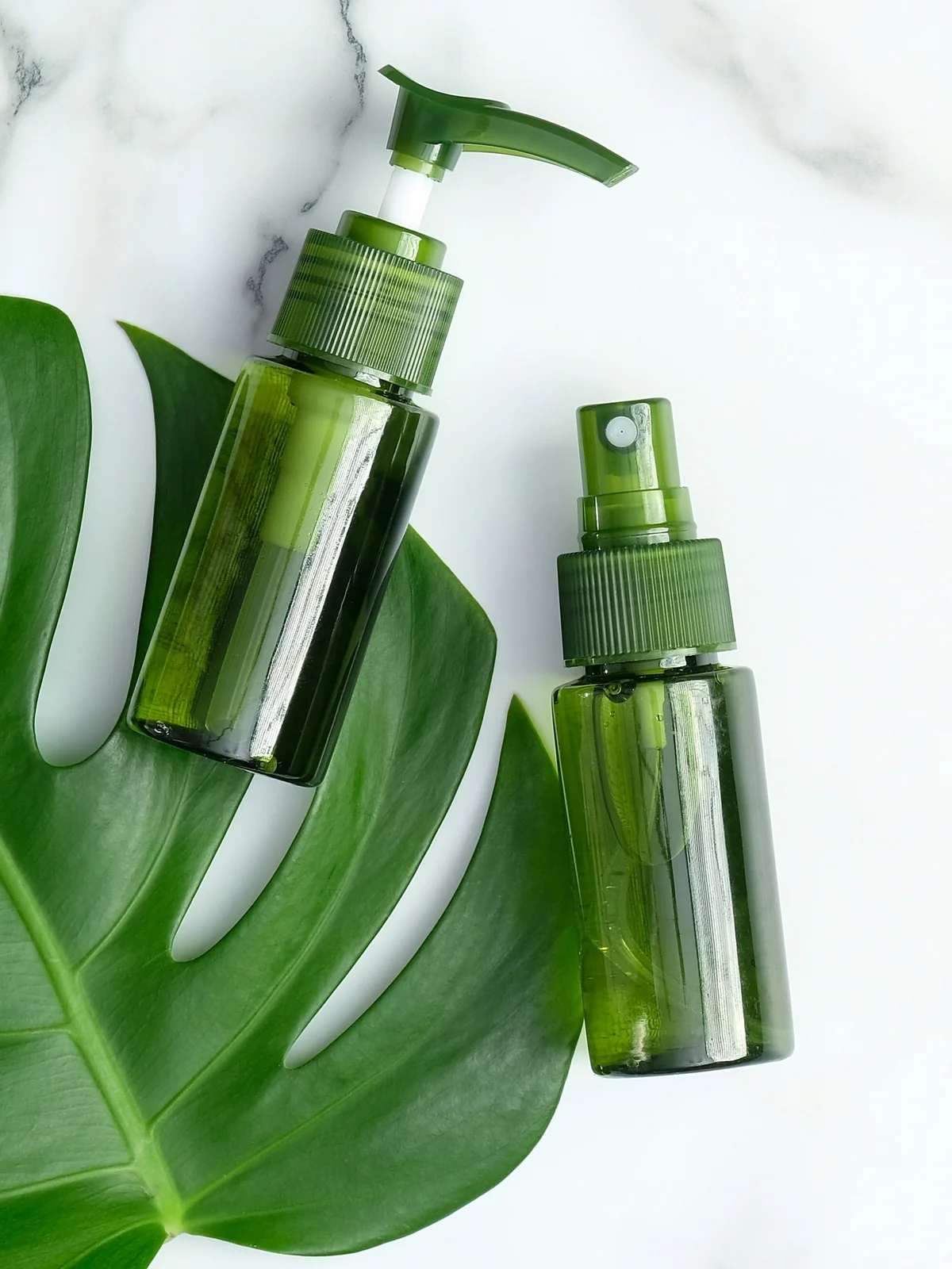 Flat lay of two green bottles of cooling gels and serums laying on a green leaf on a white marble background