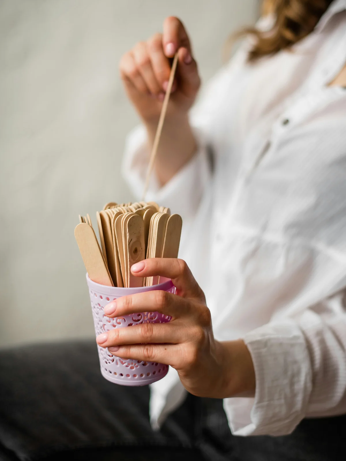Close up of a woman wearing a white shirt holding a pink pot of wooden spatulas with one hand and pulling a spatula out of the pot with the other hand