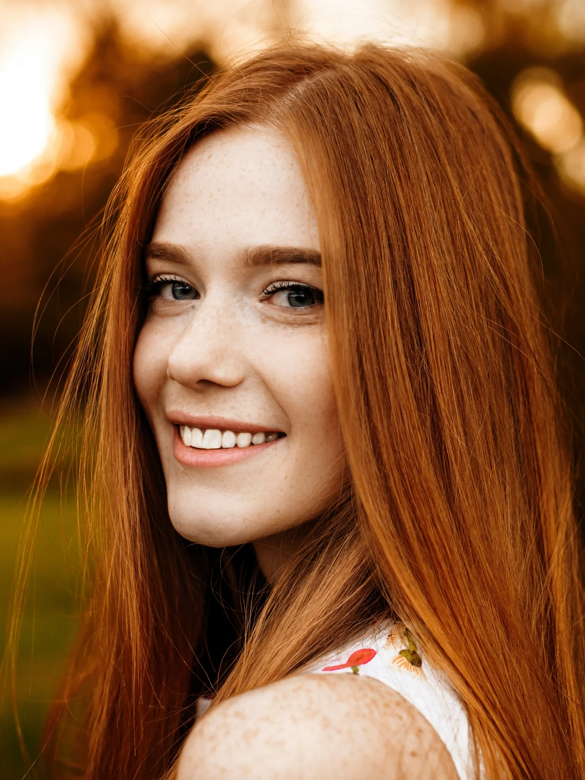 A woman with long red hair and neat, defined brows smiles as she walks outside in nature in the fading daylight