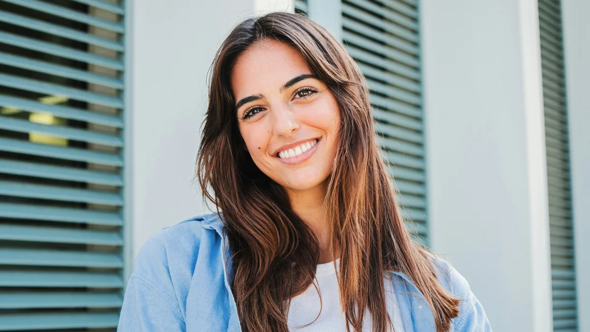 A woman with long dark hair and well-defined brows stands outside smiling in front of a building with green shutters