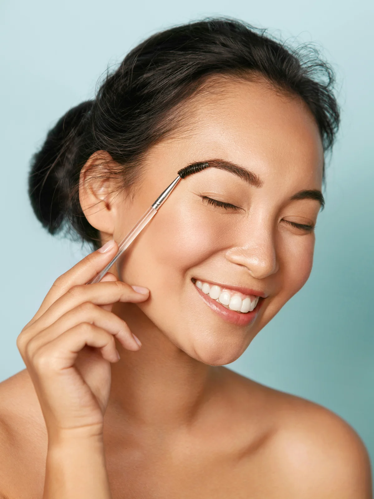 Studio shot of a woman with her hair up, smiling and neatening her eyebrows with a spoolie