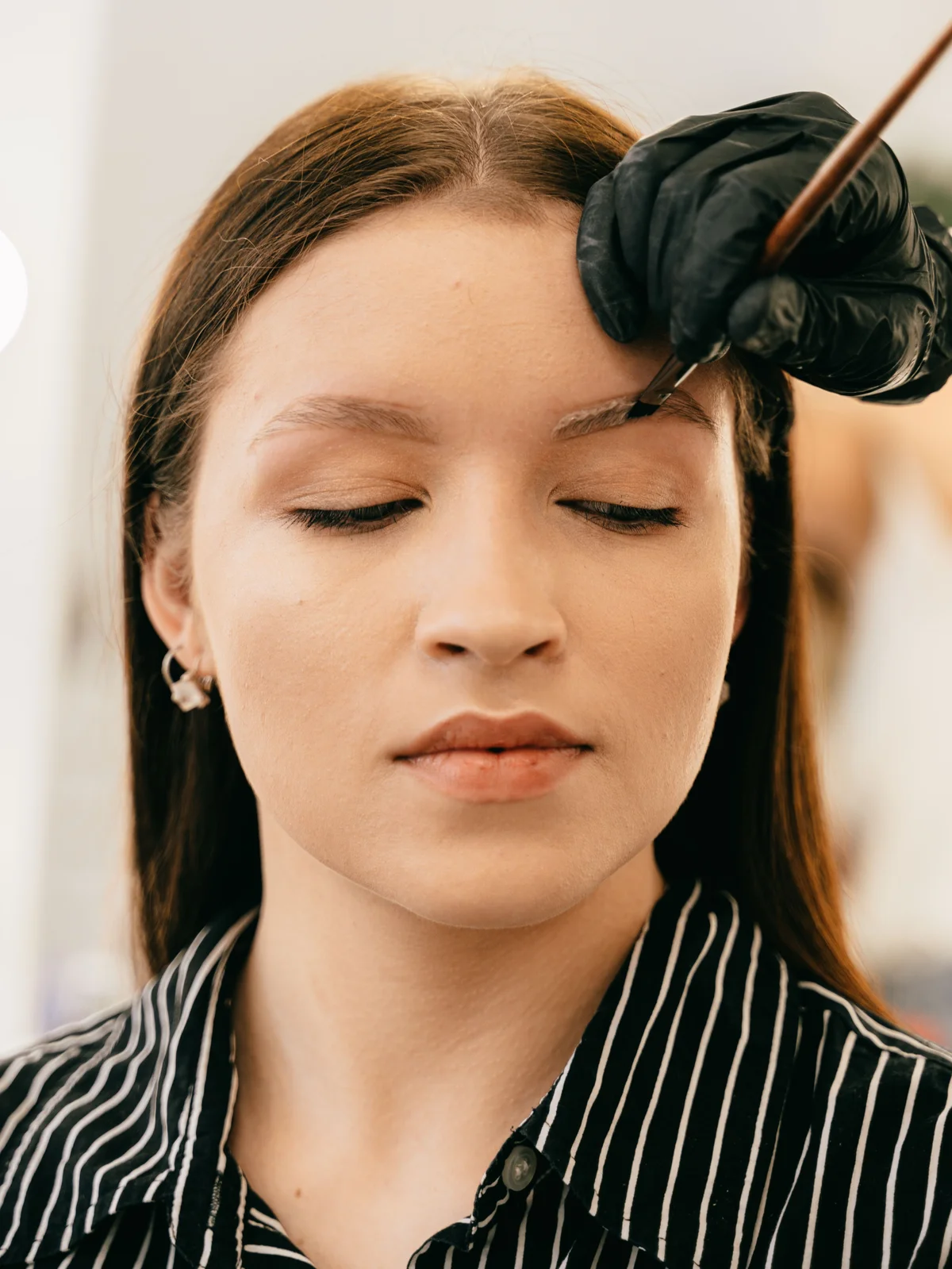 A client sits in a salon with her eyes closed as a technician carefully applies eyebrow tint with a thin brush