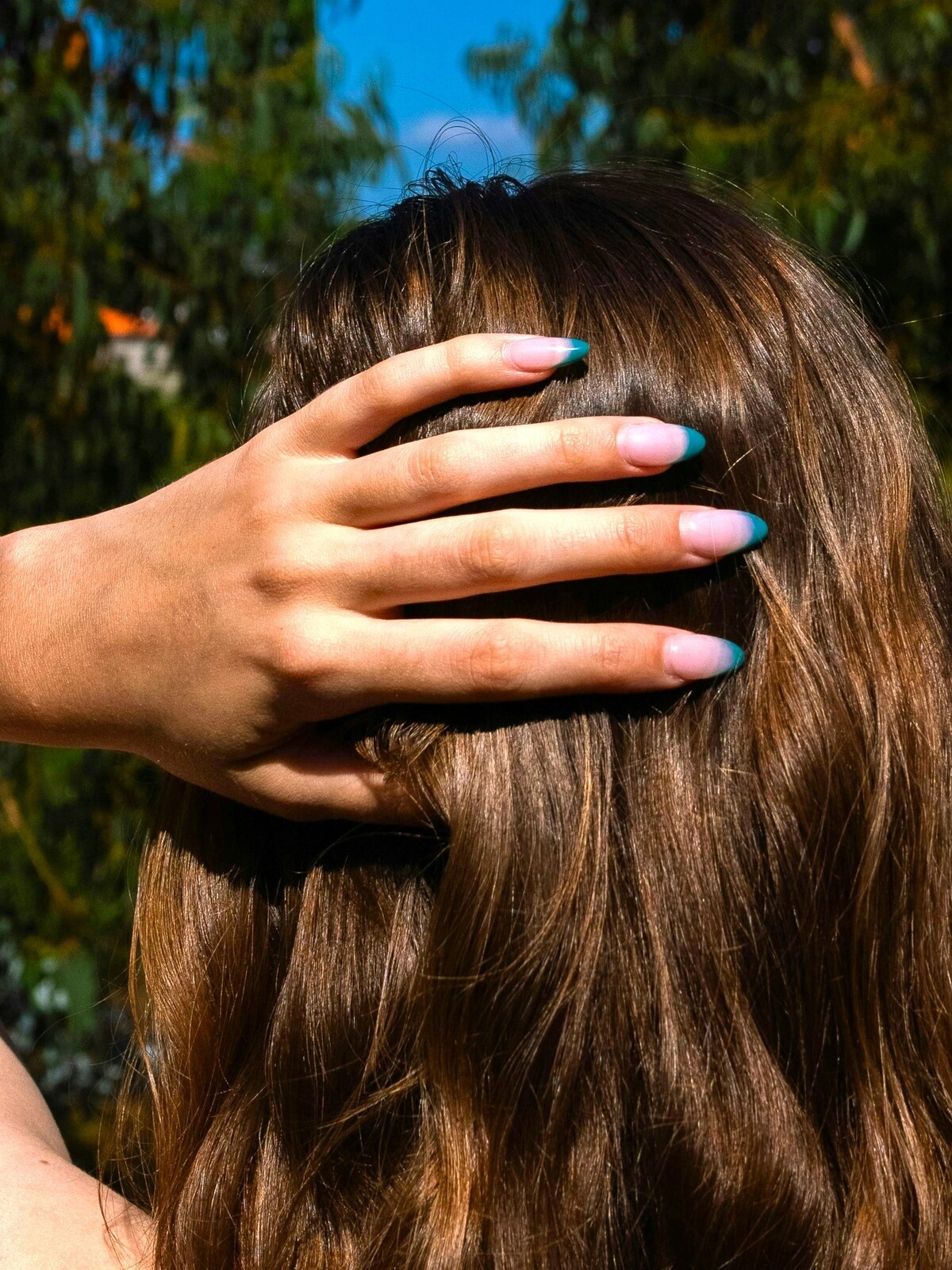 A woman’s hand rests on the back of her head showing a French tips manicure with a pale pink base and teal tips on almond nails