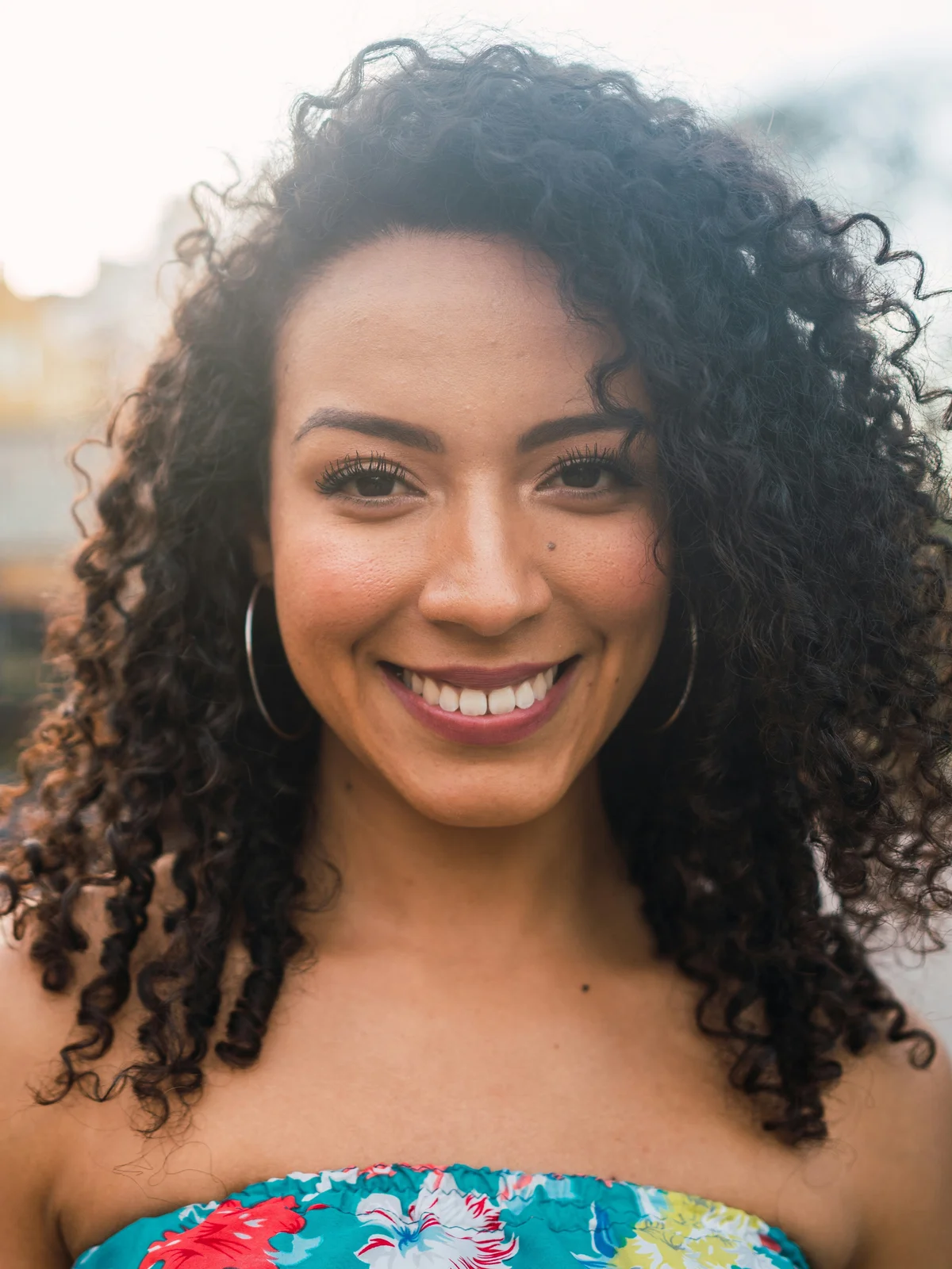 A woman with neatly groomed brows smiles at the camera wearing a floral strapless top outside in the evening sun