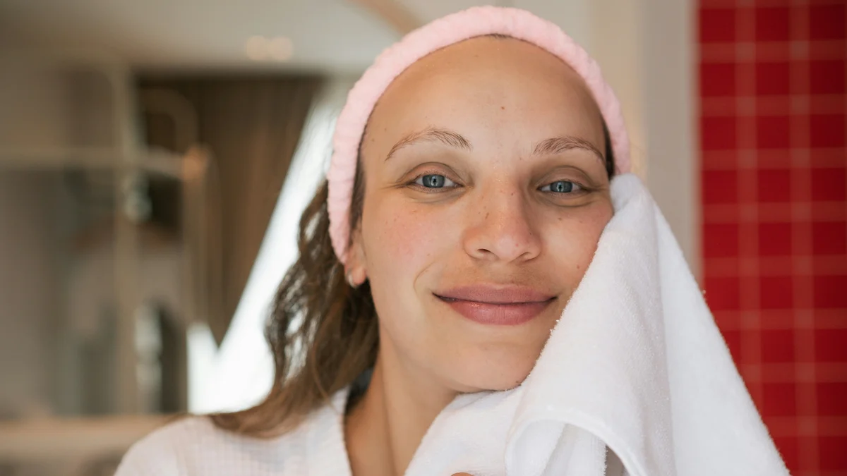 A woman with clear, glowing skin smiles at the camera wearing a pink beauty headband and patting her clean face dry with a white towel