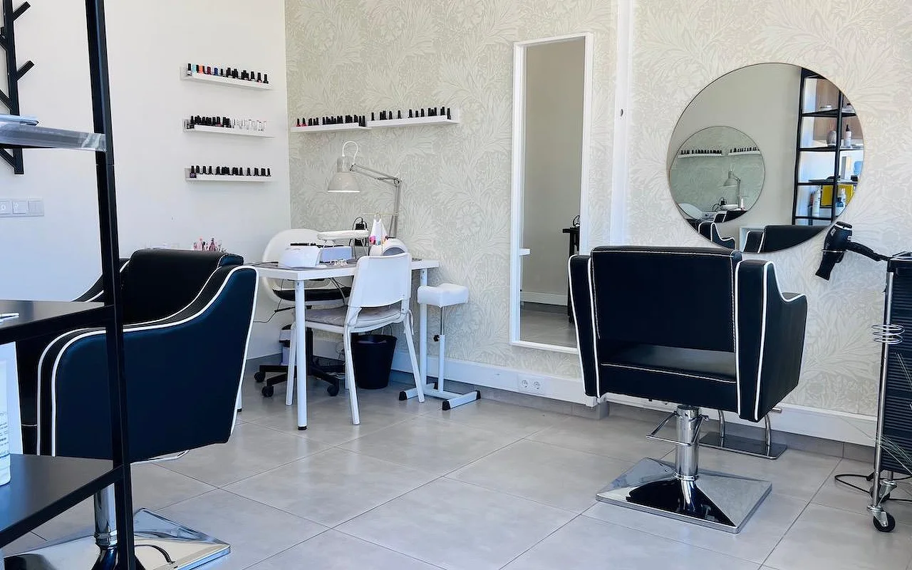 A compact hair and nail salon with black styling chairs trimmed in white piping, a round mirror and a tall mirror against grey floral wallpaper, and wall shelves lined with nail polishes.