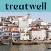 A whitewashed Algarve fishing village of terracotta-roofed houses rising up a hillside above a calm waterway, with small fishing boats moored along the quay, palm trees and a church tower under a clear blue sky.