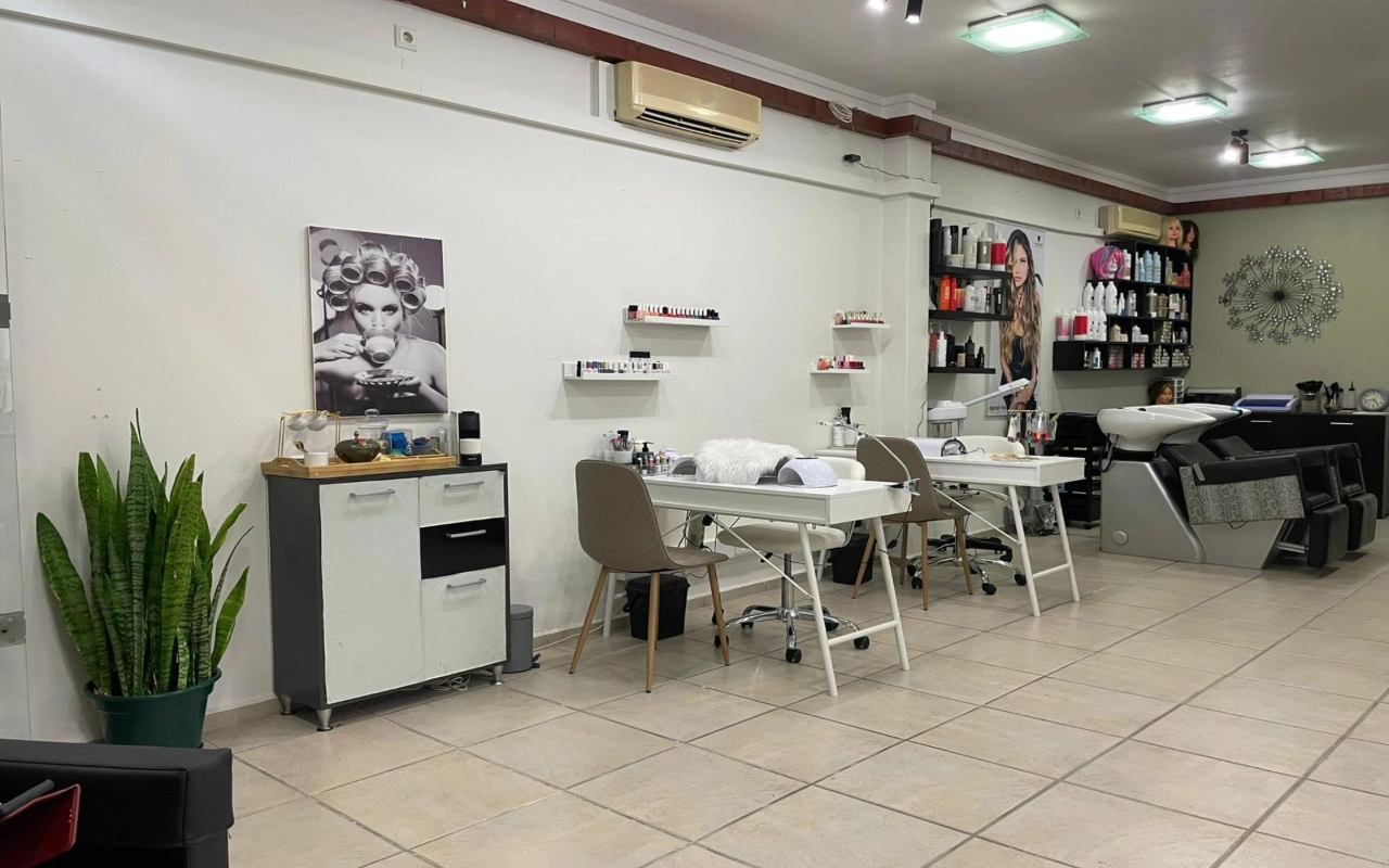 A hair and beauty salon with two white manicure tables and taupe chairs, a backwash basin and product shelves along the wall, a tall snake plant in the corner, and a large black-and-white photograph of a woman in hair curlers.