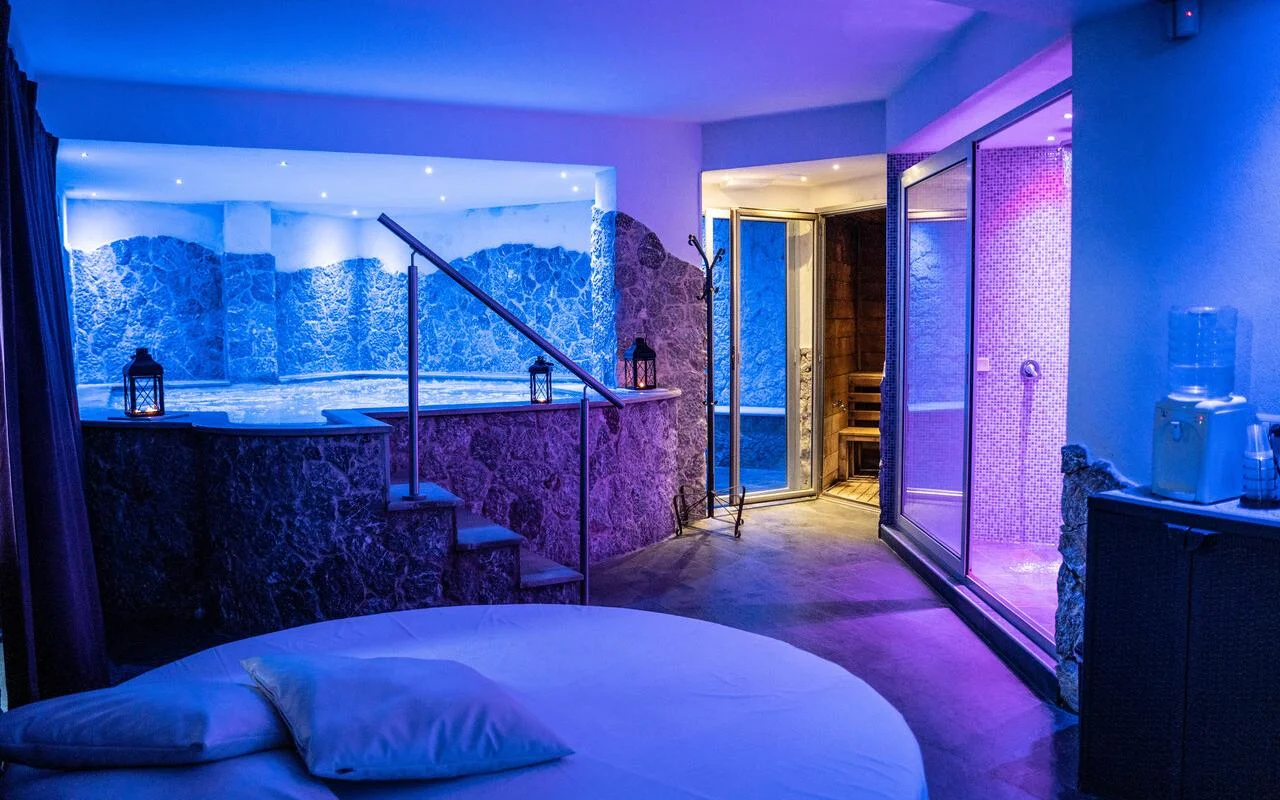 A private spa suite at Bellezza e Benessere in Taormina, lit in blue and purple, with a sunken hot tub in a stone-clad surround, a wooden sauna and a mosaic-tiled steam room behind, and a round bed in the foreground.