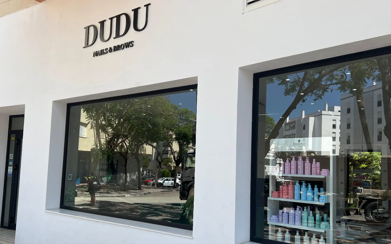 The white shopfront of Dudu Nails and Brows in Estepona, its name in dark lettering above wide windows displaying rows of haircare products.