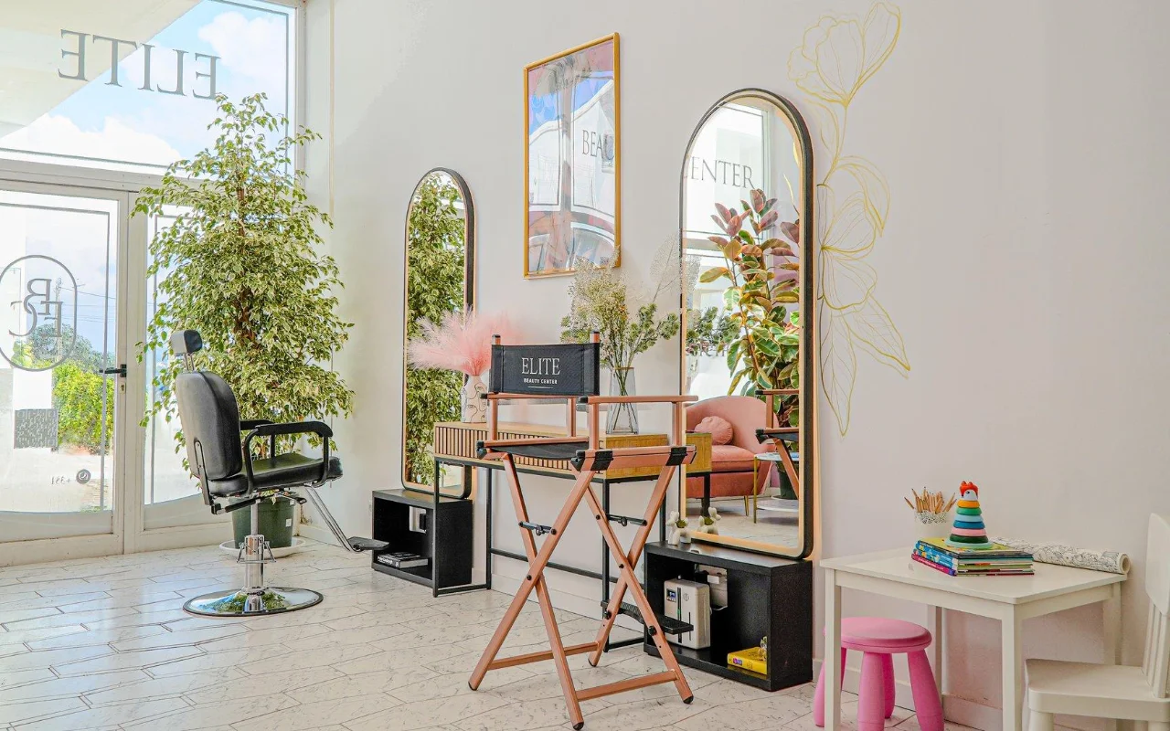 A bright, airy white beauty salon with arched gold-framed mirrors, a black styling chair and a folding director's chair, tall green plants, pink accents and a small children's corner by the window.
