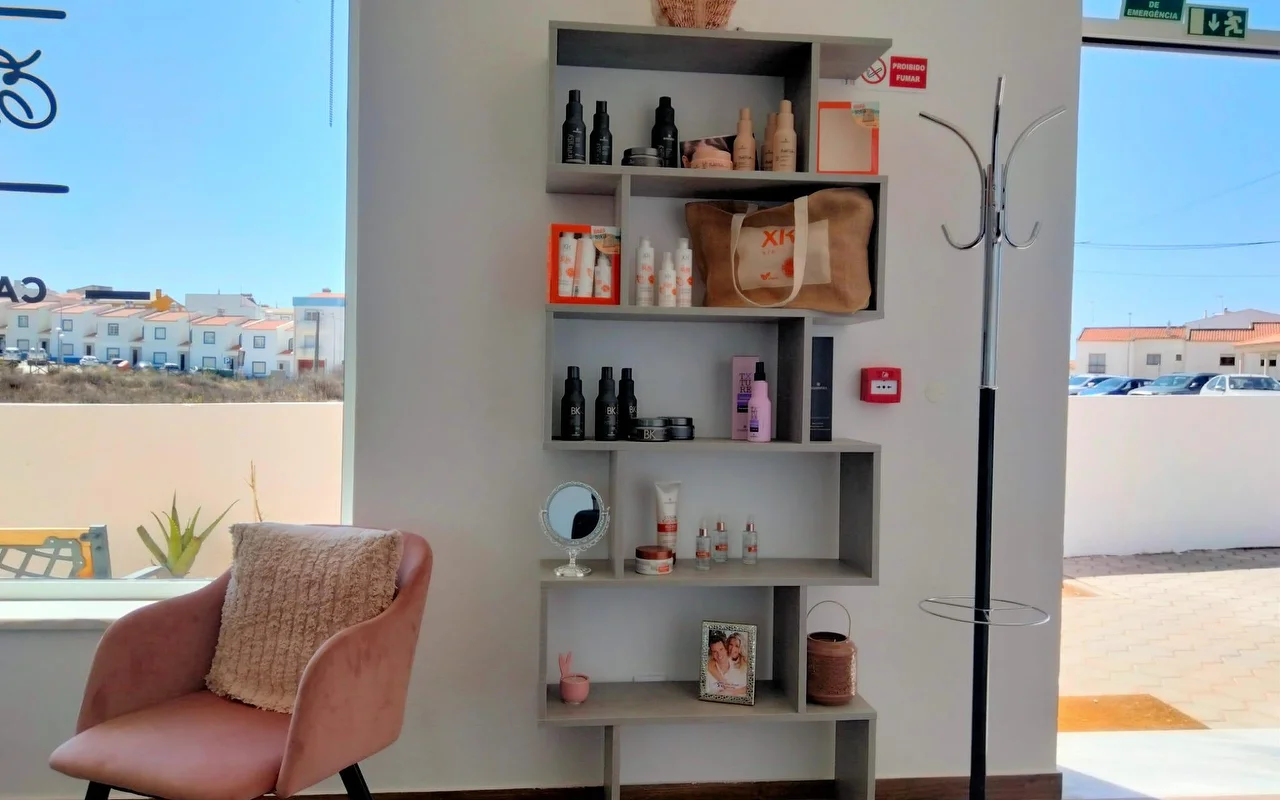 A small salon corner beside a large window, with a stepped grey shelf of hair products and a framed photo, a dusky-pink velvet armchair with a cushion, and a view out to low whitewashed houses under a blue sky.
