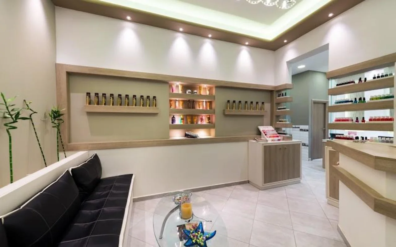 The cream-toned reception at Fabulous Nails and More in Fira, Santorini, with a black leather bench, a glass table, backlit shelves of beauty products and soft downlighting.