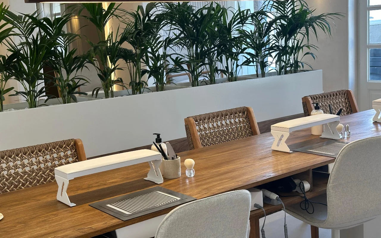 row of wooden manicure tables at Her Paros in Naoussa, each with a white arm rest and a woven rattan chair, set against tall green palms and a mirrored wall.