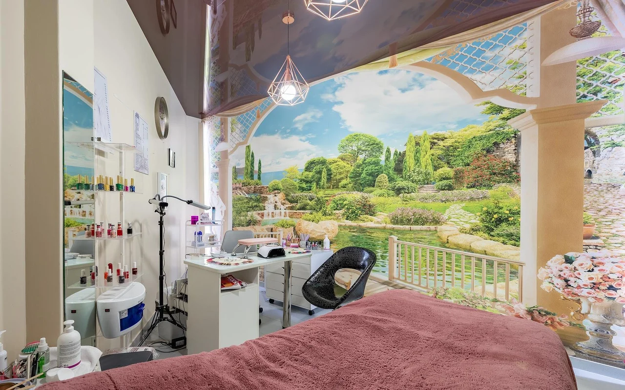 A beauty treatment room with a pink-draped bed and a white nail station, set against a large garden-and-lake wall mural, with glass shelves of products alongside.