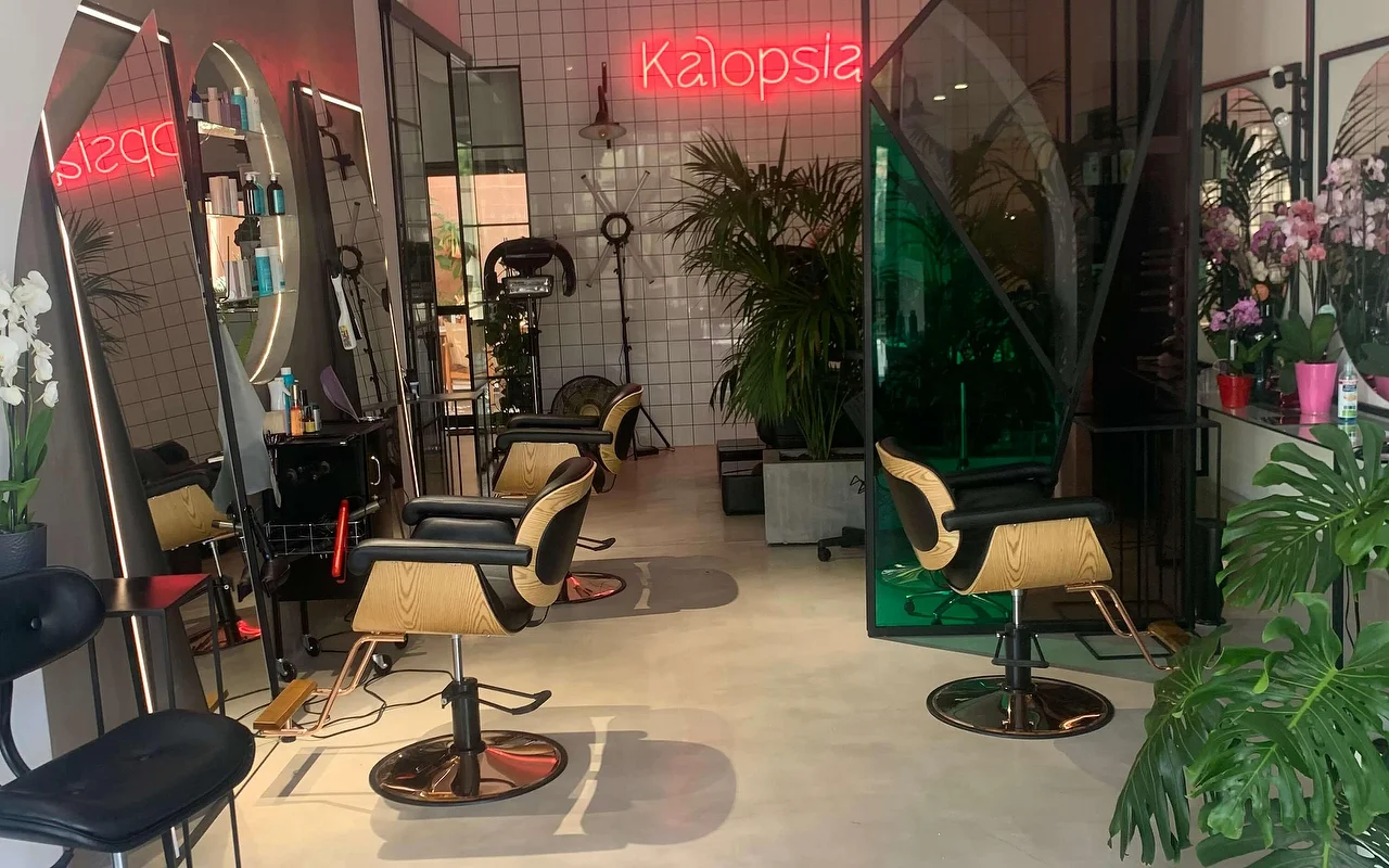 The styling floor at Kalopsia hair salon in Ermoupoli, Syros, with pale-wood and black chairs, arched backlit mirrors, leafy plants and a pink neon sign reading Kalopsia.