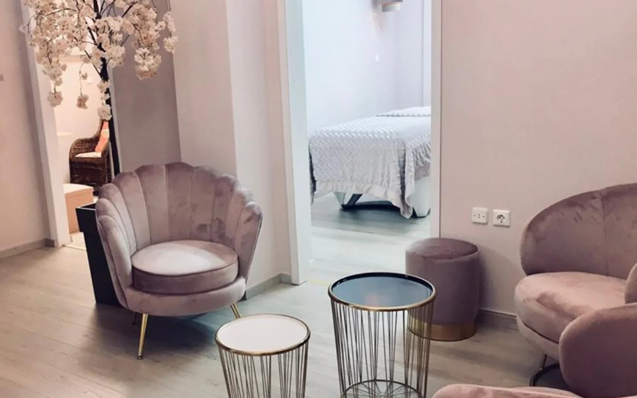 The blush-pink waiting area at Nomikou Antonia Aesthetic Lab in Syros, with a velvet scalloped armchair, slim gold side tables, a cherry-blossom branch and a treatment room beyond the doorway.