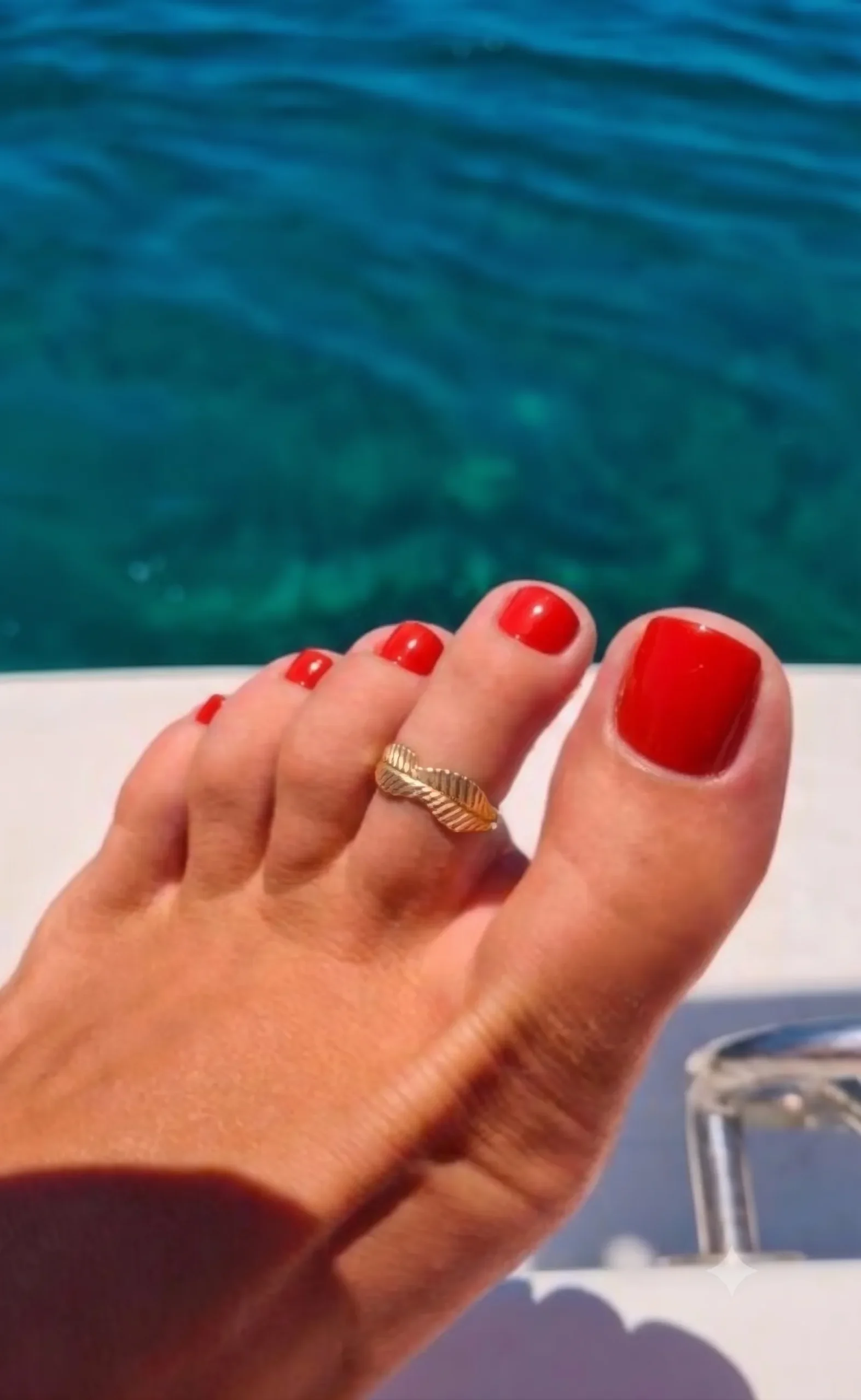 Glossy tomato red pedicure on sun-kissed foot with gold toe ring, turquoise sea in the background
