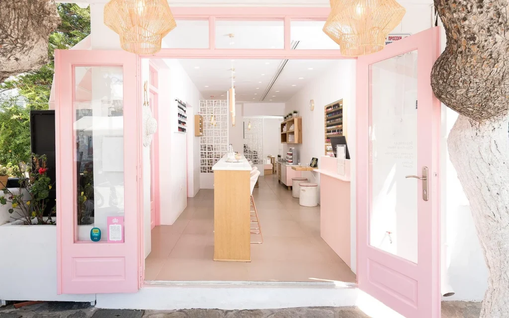 The pink-framed entrance to Vivian Nail Boutique in Naoussa, Paros, with open doors, woven pendant lights and a wooden counter, set into a whitewashed Cycladic building beside a tree.