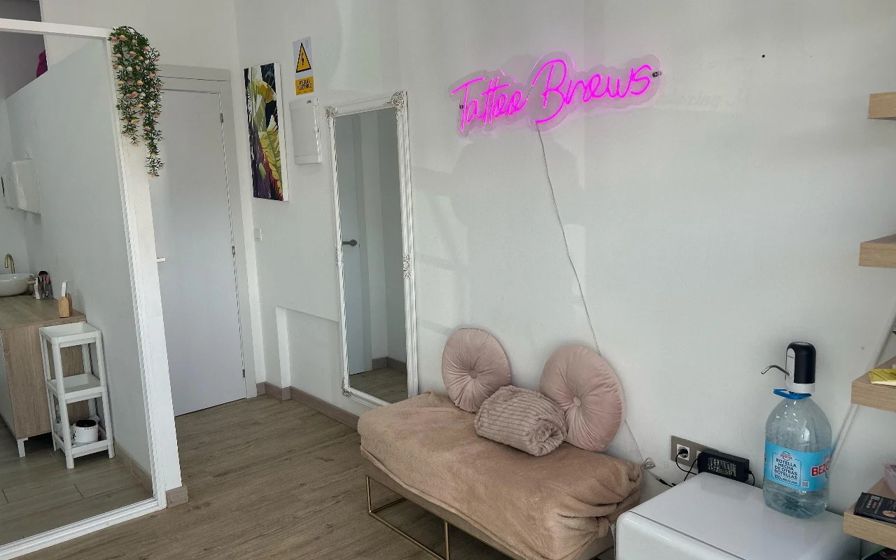 A treatment space at YG Beauty Estudio in Torremolinos, with a pink 'Tattoo Brows' neon sign, a full-length mirror and a cushioned beige daybed.